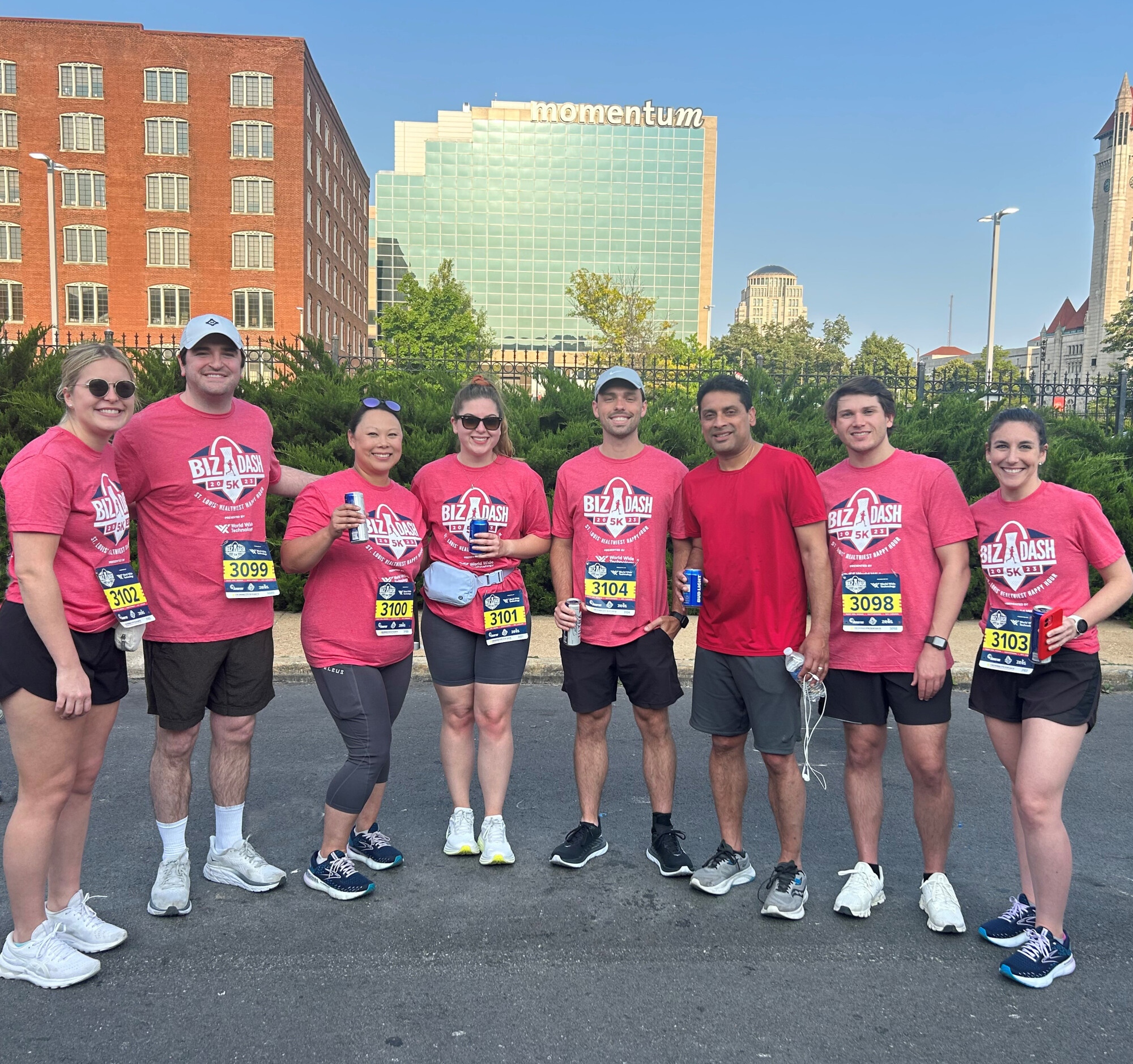 8 people in pink t-shirts, dark shorts, and running shoes with large buildings in background