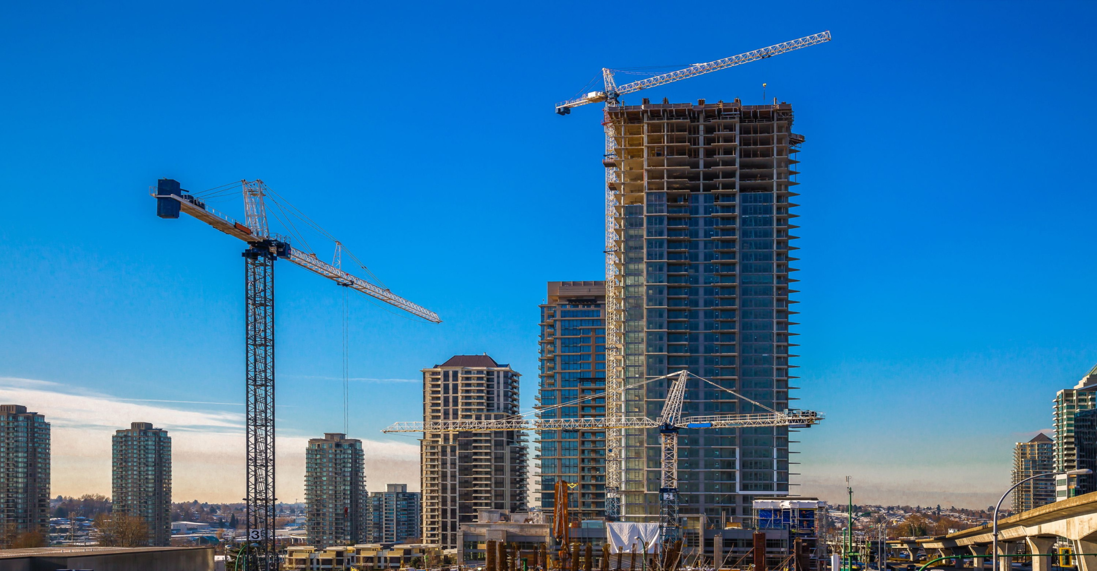 city skyline with construction scaffolding on top of skyscrapers and large cranes in foreground.