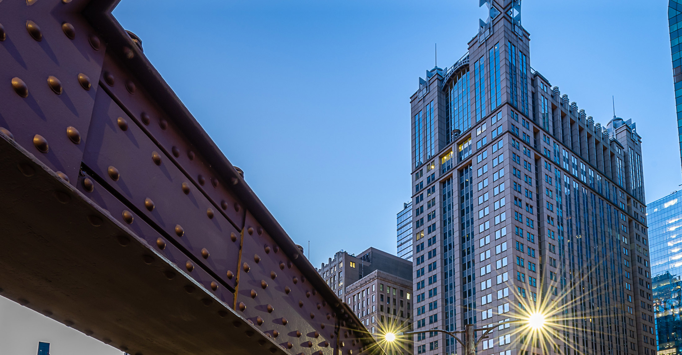 a horizontal steel girder in left foreground from a Chicago iron bridge withs skyscrapers in background on right and 2 rays of sunlight reflecting off building windows