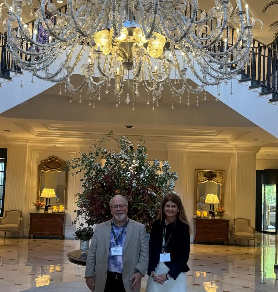 man and woman in a hotel lobby beneath a large crystal chandelier and in front of a floral arrangement.